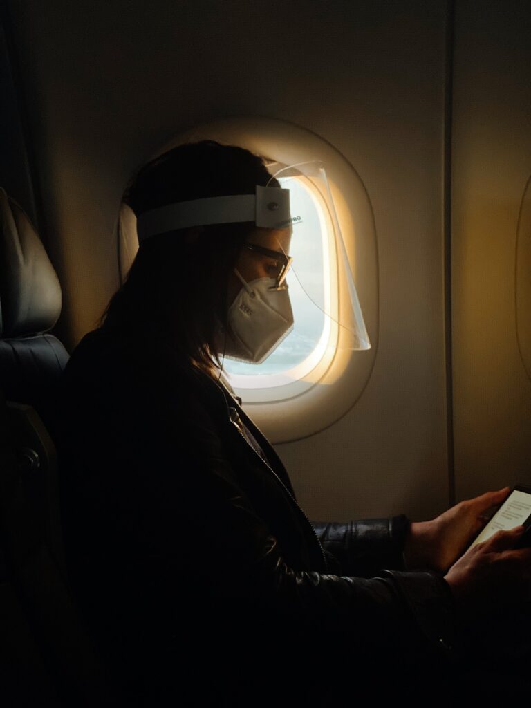 A lady sitting by the window in the flight reading a book on kindle while wearing the face mask. Travel during Covid 19 Pandemic