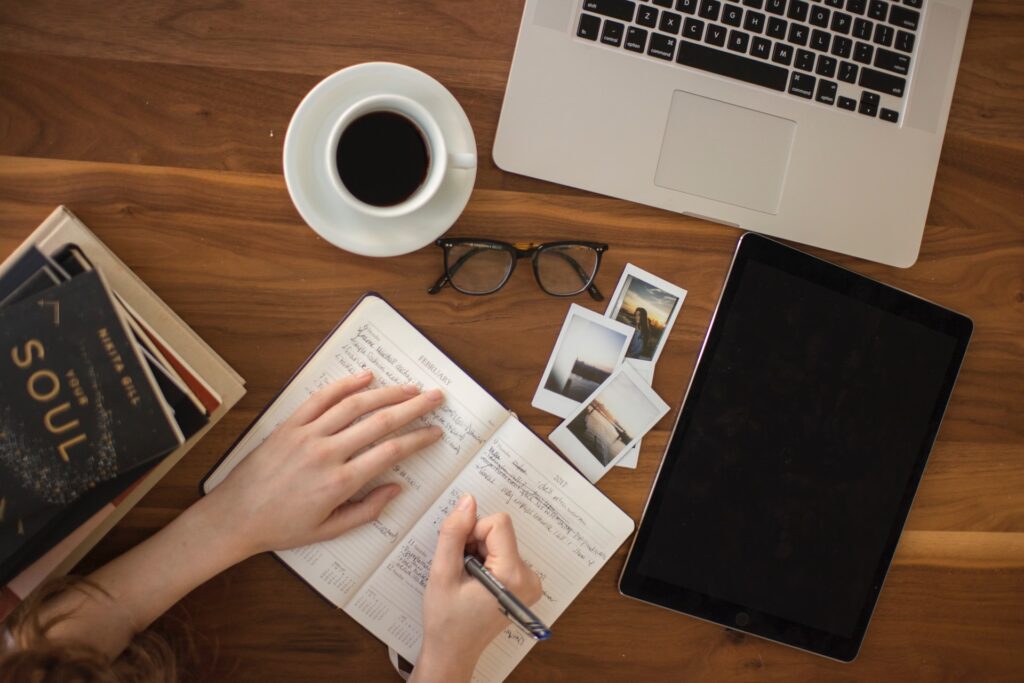 A laptop, a cup of coffee, spectacles photos, books and a person writing a diary on the table